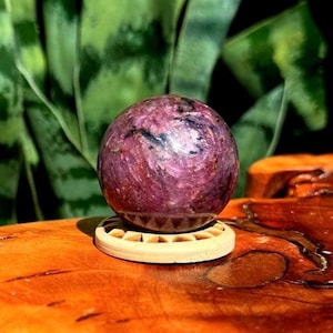 Pode incluir: Uma pedra preciosa esférica polida com uma superfície marmoreada em magenta e roxo profundo com inclusões escuras. A esfera repousa sobre um suporte circular de madeira clara com um padrão triangular esculpido ao longo da borda.