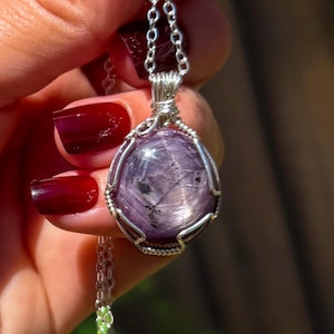 Op de afbeelding: Een ronde, gepolijste paarse edelsteen met donkere insluitingen, gevat in een zilverkleurige draadomwikkeling. De hanger hangt aan een fijne zilverkleurige ketting en is voorzien van een stevig oogje dat verbonden is met het metalen frame.