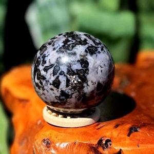 May include: A spherical decorative stone with a speckled black and white pattern. The stone is resting on a small, round, light-colored wooden stand. The background is blurred, with green foliage and a wooden surface.