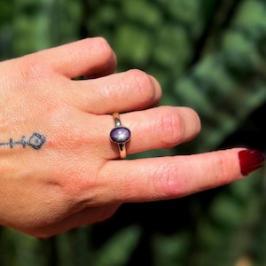 May include: A gold ring with a dark purple oval gemstone displaying a star pattern. The ring is worn on a hand with a tattoo and dark red nail varnish.
