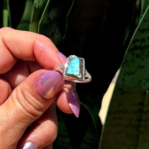 May include: A silver ring featuring a rectangular, iridescent blue gemstone. The ring has a simple band with a split design on the sides. The gemstone has a textured surface and reflects shades of blue and green.