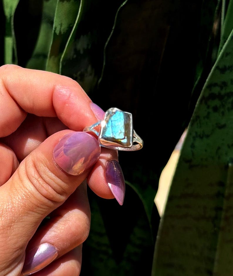 May include: A silver ring featuring a rectangular, iridescent blue gemstone. The ring has a simple band with a split design on the sides. The gemstone has a textured surface and reflects shades of blue and green.