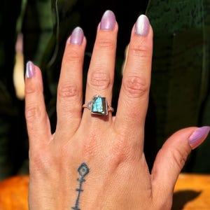 May include: A silver ring with a rectangular, iridescent blue-green stone. The ring is set on a hand with a tattoo on the wrist and light purple nail polish. The background is blurred green foliage.