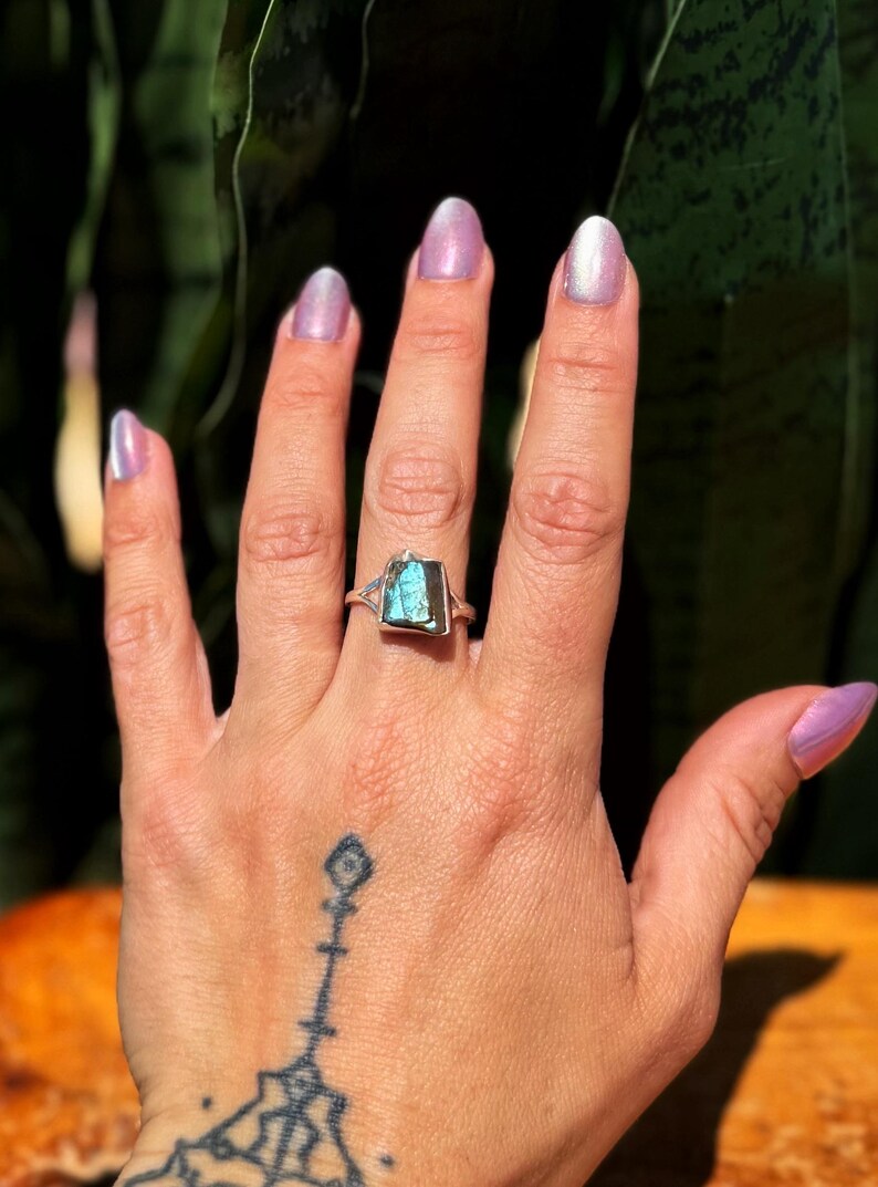 May include: A silver ring with a rectangular, iridescent blue-green stone. The ring is set on a hand with a tattoo on the wrist and light purple nail polish. The background is blurred green foliage.