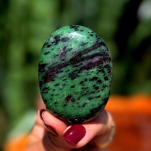 May include: Oval-shaped zoisite stone with a polished surface. The stone features a vibrant green base color with black and maroon veining throughout. The stone is held in a hand with maroon nail polish.