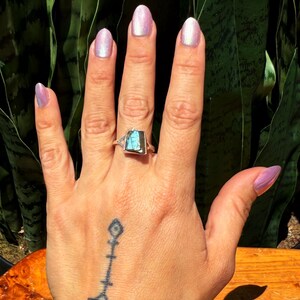 May include: A silver ring with a rectangular, iridescent blue-green gemstone. The ring is on a hand with a tattoo and light purple nail polish. The background features green foliage.