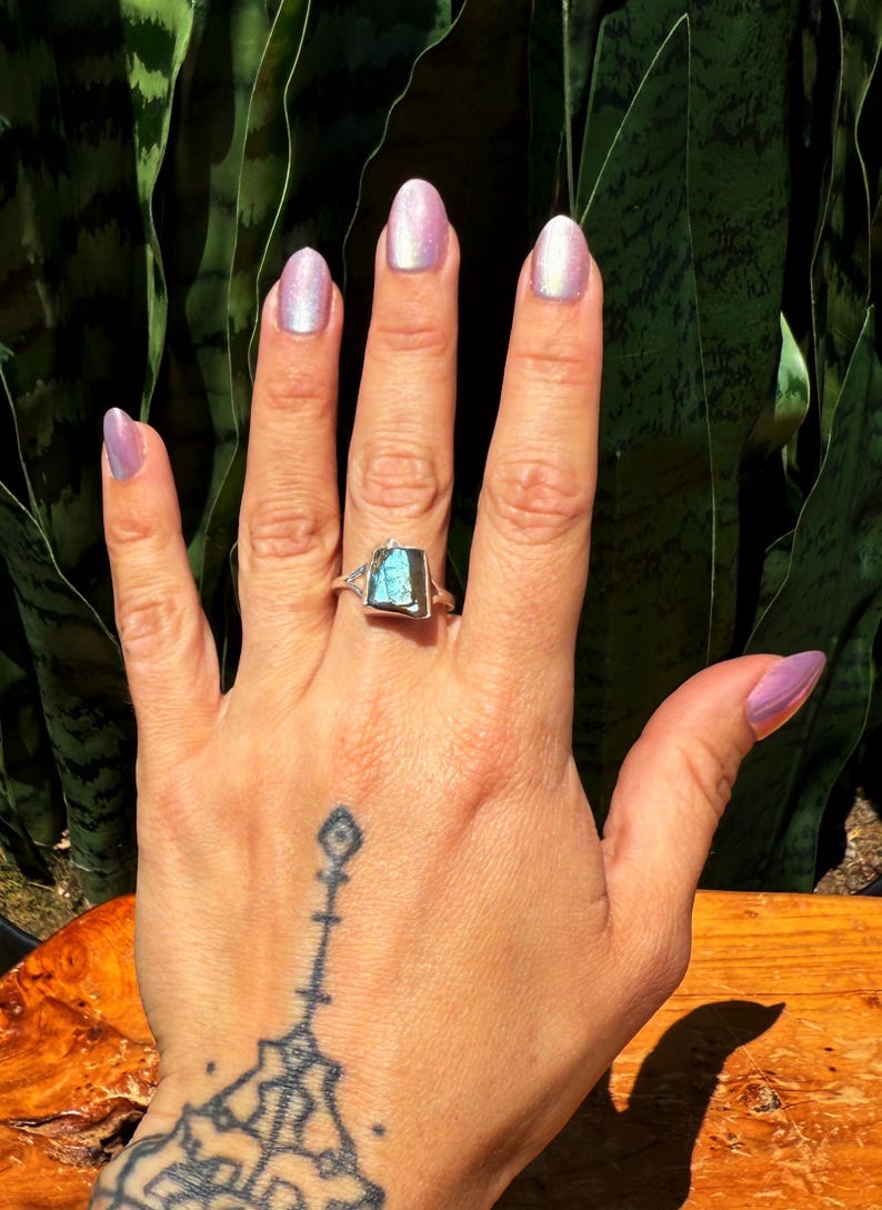 May include: A silver ring with a rectangular, iridescent blue-green gemstone. The ring is on a hand with a tattoo and light purple nail polish. The background features green foliage.