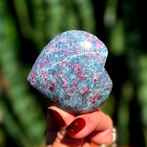 May include: A heart-shaped polished stone featuring a mottled blue-grey base color with embedded vibrant magenta and deep red crystalline inclusions throughout the surface, showcasing a smooth texture and a rounded, symmetrical heart silhouette.