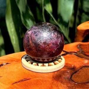 Può includere: Una pietra sferica lucida con colorazione screziata viola scuro e nera, caratterizzata da segni superficiali circolari concentrici. La sfera poggia su un supporto circolare in legno chiaro con una serie di piccoli intagli triangolari.