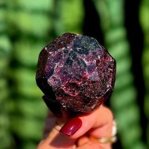 Puede incluir: Una piedra preciosa de granate facetada en color burdeos profundo y negro con una forma geométrica pulida de múltiples lados. La piedra muestra una paleta de colores oscuros y ricos con inclusiones naturales y superficies reflectantes en sus diversas caras angulares.