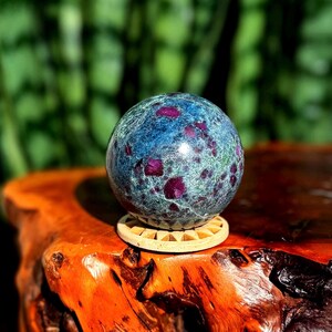 Pode incluir: Uma esfera de pedra preciosa polida, com uma cor de base azul-esverdeada e inclusões vermelhas vibrantes. A esfera repousa sobre um pequeno suporte de madeira redondo, contra um fundo verde desfocado. A esfera tem cerca de 7.6 cm de diâmetro.