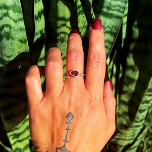 May include: A silver ring with a red gemstone set in a teardrop shape. The ring is worn on the middle finger of a hand with a tattoo.