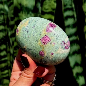 Pode incluir: Uma pedra polida de formato oval com uma cor de base verde-clara, com inclusões de cristais magenta vibrantes espalhadas e manchas escuras por toda a superfície, exibindo uma textura lisa e arredondada e padrões minerais naturais.