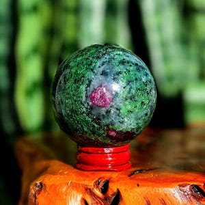 May include: A polished gemstone sphere, primarily green with red inclusions, rests on a vibrant red stand. The sphere's surface has a speckled appearance. The stand sits on a wooden surface, with a blurred green background.