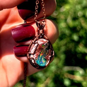 May include: A wire-wrapped pendant necklace featuring a central oval-shaped stone with a textured, reddish-brown matrix and a vibrant, iridescent play-of-color center displaying flashes of green, blue, and orange, suspended on a copper-toned chain.