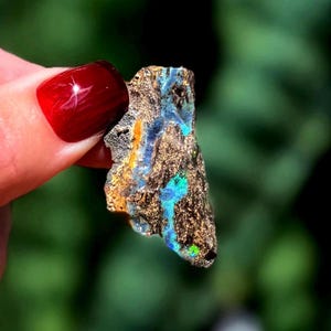May include: A rough-cut opal gemstone with a dark brown matrix, displaying vibrant flashes of blue, green, and orange. The gemstone is held up against a blurred green background, showcasing its natural beauty and unique color play.