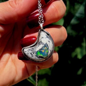 May include: A crescent moon-shaped pendant necklace featuring a white stone with black veining and a central iridescent inlay displaying flashes of green, orange, and blue. The pendant is set in a silver-toned metal frame with a beaded edge.