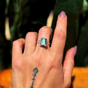 May include: A silver ring with a rectangular, blue-green gemstone. The ring is on a hand with a tattoo on the wrist. The gemstone is approximately 0.75 inches long and 0.5 inches wide.