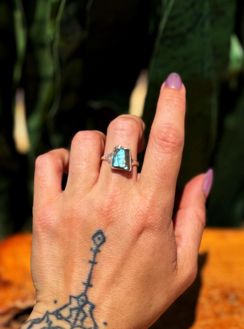 May include: A silver ring with a rectangular, blue-green gemstone. The ring is on a hand with a tattoo on the wrist. The gemstone is approximately 0.75 inches long and 0.5 inches wide.