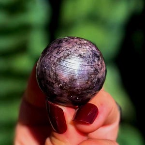 May include: A polished spherical gemstone featuring a deep purple and plum base color with intricate black mineral inclusions and a distinct chatoyant sheen that creates a horizontal light reflection across the rounded surface of the stone.