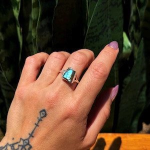 May include: A silver ring with a rectangular, iridescent blue gemstone. The ring is set on a hand with a tattoo on the wrist and purple nail polish. The background is a blurred view of green foliage.