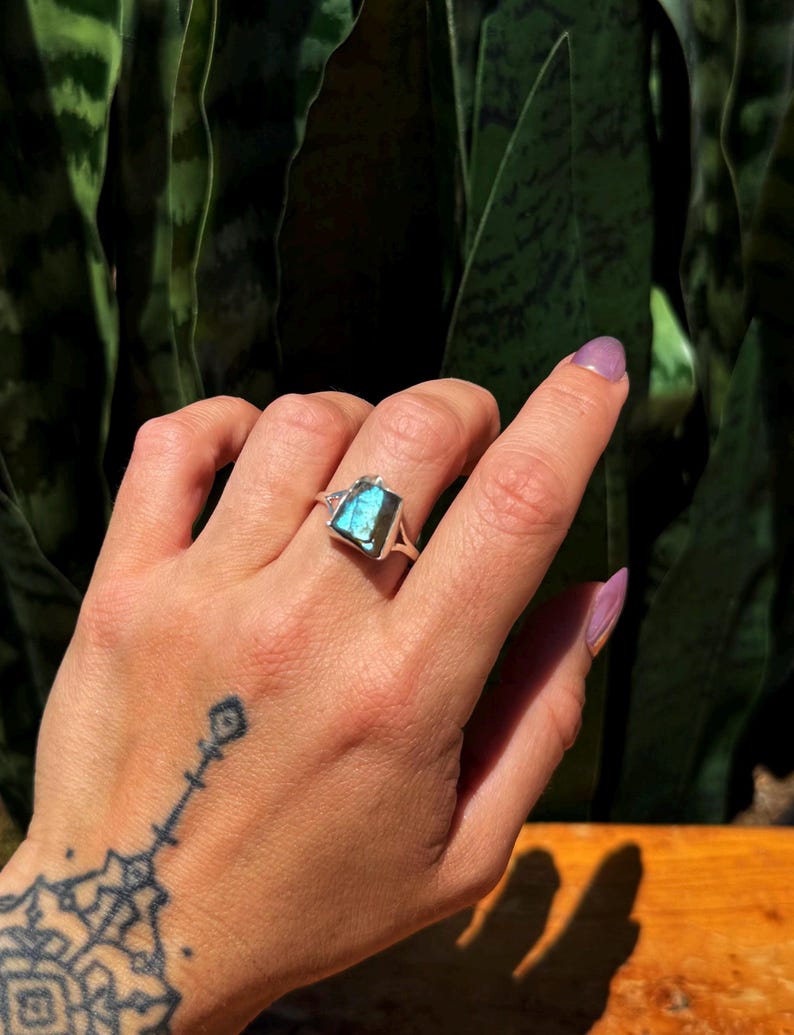 May include: A silver ring with a rectangular, iridescent blue gemstone. The ring is set on a hand with a tattoo on the wrist and purple nail polish. The background is a blurred view of green foliage.