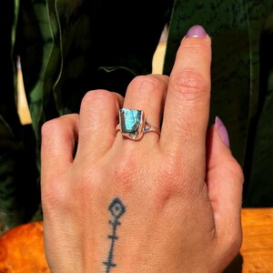 May include: A silver ring featuring a rectangular, iridescent blue-green gemstone. The ring's band has a simple design, and the gemstone is the focal point. The ring is displayed on a hand with a tattoo.