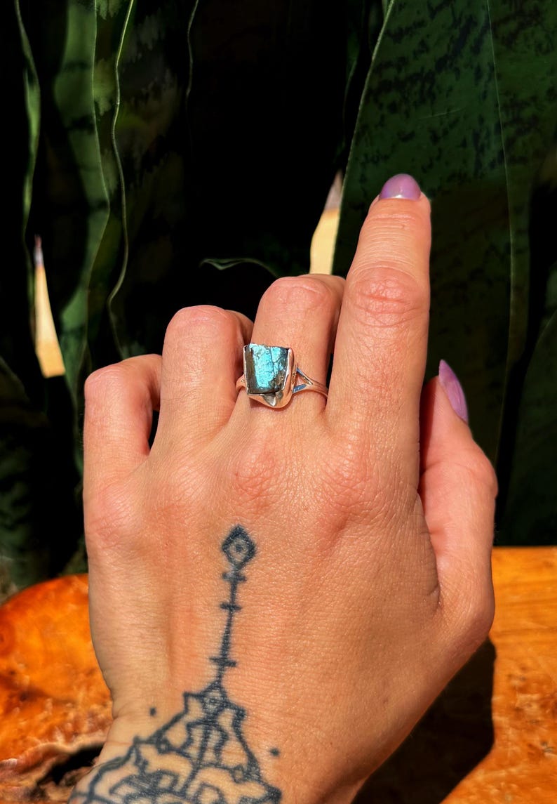May include: A silver ring featuring a rectangular, iridescent blue-green gemstone. The ring's band has a simple design, and the gemstone is the focal point. The ring is displayed on a hand with a tattoo.