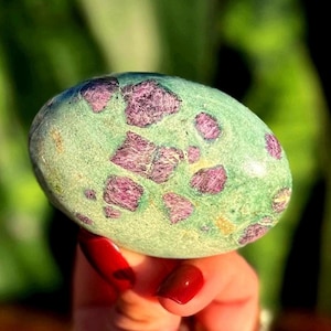 May include: An oval-shaped stone with a light green base and irregular patches of deep pink and purple. The stone is held in a hand with red nail polish, against a blurred green background. The stone's surface appears smooth and polished.