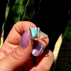 May include: A silver ring featuring a rectangular, raw-cut, blue-green gemstone. The ring is held against a backdrop of green foliage and a dark background, highlighting the stone's color and texture. The ring's band has a split design.