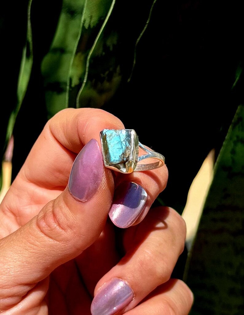 May include: A silver ring featuring a rectangular, raw-cut, blue-green gemstone. The ring is held against a backdrop of green foliage and a dark background, highlighting the stone's color and texture. The ring's band has a split design.