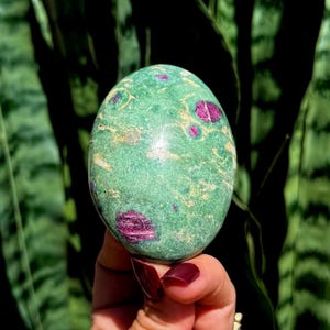 Pode incluir: Uma pedra polida de formato oval com uma base de cor verde vibrante, apresentando inclusões distintas de cor magenta profundo e veios subtis de cor clara por toda a superfície do espécimen mineral.