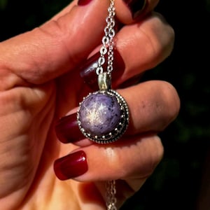 May include: A round, cabochon-cut purple star sapphire pendant set in a decorative silver bezel. The gemstone displays a distinct six-rayed star effect on its surface. The pendant hangs from a delicate silver chain with small, circular links.