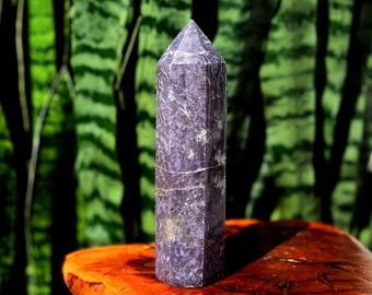 May include: A tall, purple crystal obelisk with white veining, standing on a rustic, orange-brown wooden surface. The background features blurred green foliage. The crystal has a pointed top and a polished, faceted surface.