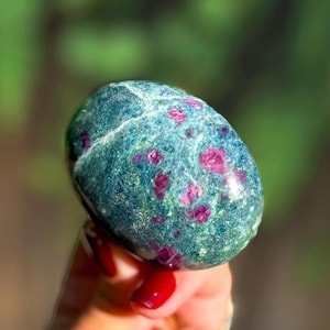 May include: A polished, oval-shaped gemstone featuring a deep teal and green base color with embedded, vibrant magenta-pink crystalline inclusions throughout the surface. The stone has a smooth, glossy texture with visible natural mineral veins.