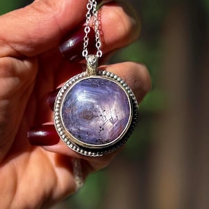 May include: A round, light purple star sapphire pendant set in a silver bezel with a beaded edge. The gemstone displays a visible six-rayed star pattern on its surface. The pendant hangs from a delicate silver chain link necklace.