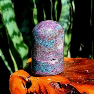May include: A polished, dome-shaped stone sculpture featuring a mottled texture with vibrant swirls of deep magenta, bright pink, and metallic teal blue. The stone has a smooth, rounded top and a small, natural crystalline indentation on its side.