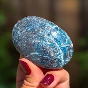 May include: A polished, oval-shaped blue apatite gemstone with a smooth, glossy surface. The stone displays deep shades of blue with visible internal crystalline structures, white mineral inclusions, and dark flecks throughout its dense texture.