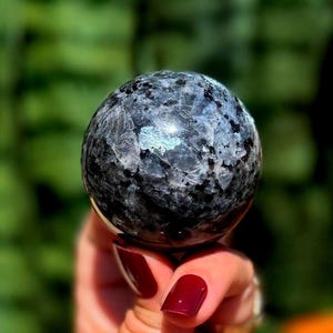 May include: A speckled, spherical gemstone in shades of black and grey, held in a hand with red nail polish. The sphere has a reflective, iridescent quality, suggesting a crystalline structure. The background is a blurred green.
