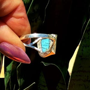 May include: A silver ring with a rectangular turquoise stone. The ring has a unique, open design with a polished finish. The turquoise stone is a vibrant blue-green color, set in a silver bezel.