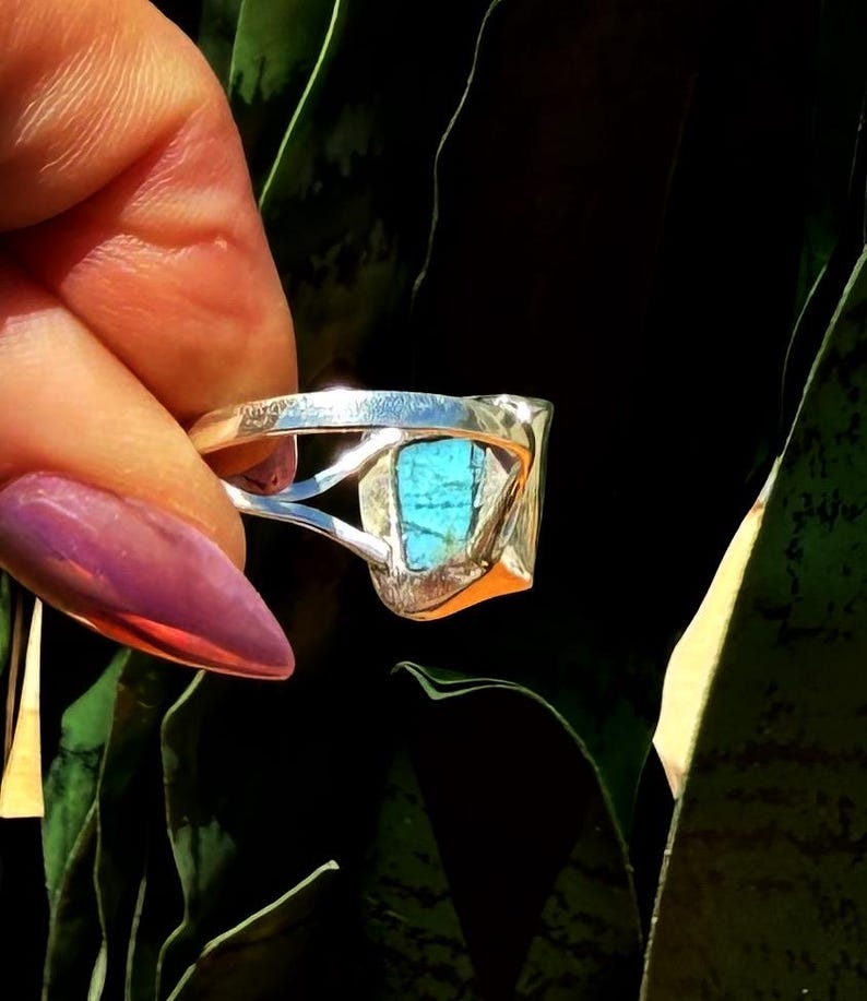 May include: A silver ring with a rectangular turquoise stone. The ring has a unique, open design with a polished finish. The turquoise stone is a vibrant blue-green color, set in a silver bezel.