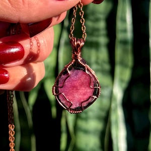 May include: A copper-colored pendant necklace featuring a pink gemstone. The gemstone is encased in a wire-wrapped setting and hangs from a delicate chain. The pendant has a teardrop shape and is held up by a hand with red nail polish.