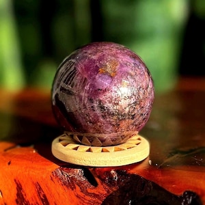 May include: A polished, spherical gemstone in shades of deep purple, maroon, and brown, resting on a small, round wooden stand. The gemstone has a reflective surface and sits on a wooden stand with a diameter of approximately 2 inches.