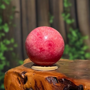 Op de afbeelding: Een gepolijste, bolvormige roze edelsteen met een kristallijne, gevlekte textuur. De bol rust op een kleine, ronde, lichtgekleurde houten standaard. Het oppervlak van de steen vertoont verschillende tinten dieproze en lichter roze met een gladde afwerking.