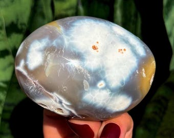 May include: A polished, oval-shaped stone with a marbled pattern of white, gray, and blue hues. The stone has small, reddish-brown spots and a smooth, rounded surface. The background is a blurred green.