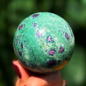 Pode incluir: Uma esfera verde com manchas vermelhas escuras. A pedra preciosa é segurada em uma mão com esmalte de unhas bordô. A esfera tem uma superfície texturizada e é provavelmente um item decorativo ou um cristal.