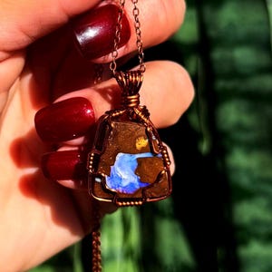 May include: A copper-colored pendant necklace featuring a triangular, raw stone with blue and brown hues. The stone is encased in a wire-wrapped setting and hangs from a delicate chain. The necklace is held up by a hand with red nail polish.