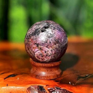 May include: A spherical gemstone, predominantly purple with black inclusions, displayed on a small, turned wooden stand. The gemstone sits on a wooden surface, with a blurred green background. The stand is made of wood.