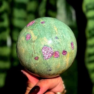May include: A green sphere with red and pink inclusions. The polished stone is held in a hand with a dark red manicure. The background is a blurred green.