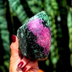 Pode incluir: Um espécime mineral de zoisite bruto com uma inclusão proeminente de cristal de rubi rosa magenta vibrante, incrustada numa matriz de zoisite verde floresta escura e preta, exibindo formações cristalinas naturais e contraste de cores.